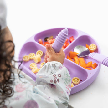 A child enjoys colorful foods with a purple silicone spoon, divided plate, and Bumkins Floral sleeved bib made of waterproof fabric.