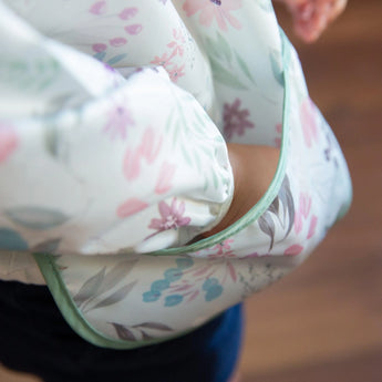 Close-up of a child wearing the Bumkins Sleeved Bib: Floral, made from waterproof fabric, with their hand tucked in a pocket.