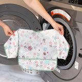 Hands hold a Bumkins Floral Sleeved Bib in waterproof fabric for baby-led weaning in front of an open washing machine.