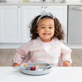 In a modern white kitchen, a young girl with curly hair and gray bow wears a Bumkins Sleeved Bib: Lace, sitting at the table with fruit.