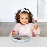 A curly-haired toddler in a Bumkins Sleeved Bib: Lace smiles, holding a fork with fruit, seated at a table of assorted foods.