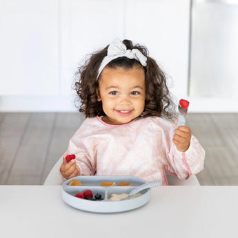 A curly-haired toddler in a Bumkins Sleeved Bib: Lace smiles, holding a fork with fruit, seated at a table of assorted foods.