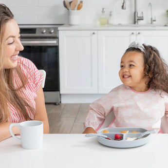 A woman and girl, using Bumkins Sleeved Bib: Lace, smile at each other over snacks and a cup in the kitchen.