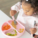 A toddler enjoys baby-led weaning with pasta, carrots, and avocado from a pink plate while using utensils and wearing a Bumkins Sleeved Bib: Lace.