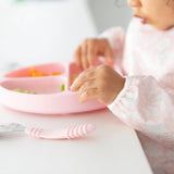 A toddler in a Bumkins Sleeved Bib: Lace eats from a sectioned plate, embracing baby-led weaning. A pink spoon is on the white table.