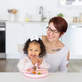 A bespectacled woman watches a child eat from a pink plate using a fork, practicing baby-led weaning, with a Bumkins Sleeved Bib.