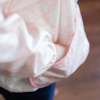 A close-up shows someone in a pink, floral waterproof Bumkins Sleeved Bib: Lace with one hand tucked into the pocket.