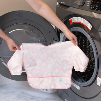 A person holds a Bumkins Sleeved Bib: Lace, a floral waterproof bib, in front of an open washing machine.