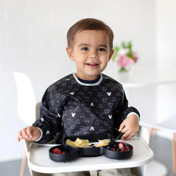 A toddler in a high chair, smiling with a divided plate of pasta and berries, wears a Bumkins Mickey Mouse Icon Black + White bib.