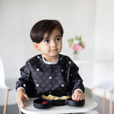 A dark-haired child in a high chair, wearing a Bumkins Sleeved Bib: Mickey Mouse Icon Black + White, eats from a black tray.