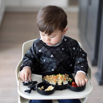 A toddler in a high chair with a Bumkins Mickey Mouse bib eats from a divided, stain-resistant plate filled with rice and fruit.