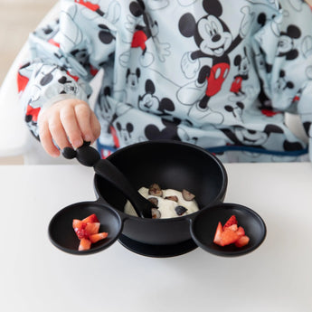 A child in a Mickey Mouse outfit enjoys strawberry yogurt, wearing a Bumkins Sleeved Bib: Mickey Mouse, at the white table.