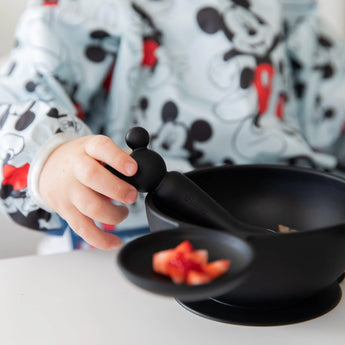 Child in a Bumkins Mickey Mouse sleeved bib holds a black spoon with Mickey handle beside a matching bowl of food.