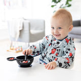 A toddler in a Bumkins Mickey Mouse sleeved bib smiles while eating from a black bowl at a white table.