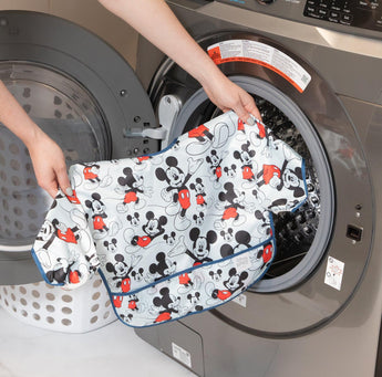 Hands placing a Disney-themed shirt into a front-loading washing machine alongside a Bumkins Sleeved Bib: Mickey Mouse.