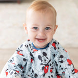 Smiling baby wearing a Bumkins Mickey Mouse sleeved bib, sitting indoors.