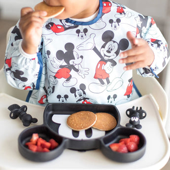 A child enjoys a pancake from a Mickey Mouse plate, wearing a Bumkins Sleeved Bib: Mickey Mouse for mealtime fun.