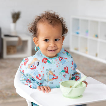 A curly-haired toddler in a high chair wears a Bumkins Sleeved Bib: Ariel with a bowl and spoon in front. Waterproof fabric for easy cleanup.