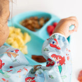 Child enjoys snacks on a blue plate, wearing a Bumkins Sleeved Bib: Ariel to catch every crumb.