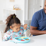 A child in a Bumkins Sleeved Bib: Ariel eats from a blue plate with a fork, sitting beside a smiling man.