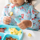 A child in a Bumkins Sleeved Bib: Ariel holds a fork with scrambled eggs by a sectioned breakfast plate at the table.