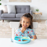 A child smiles while eating in a blue Bumkins Ariel bib with a blue sectioned plate at a white table; gray couch in the background.