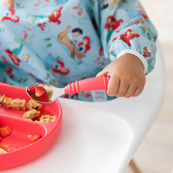 A child in a Bumkins Sleeved Bib: Ariel holds a spoon of fruit over a red plate with waffle pieces.