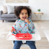 A cheerful child with curly hair, wearing a Bumkins Sleeved Bib: Ariel, smiles while holding food next to a red plate on the tray.