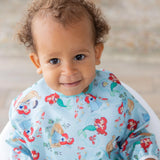 Toddler smiles at the camera wearing a colorful Bumkins Sleeved Bib: Ariel with cartoon mermaids.