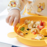 A childs hand uses a fork to eat diced fruits from a yellow plate, with Bumkins Sleeved Bib: Winnie Loves Hunny aiding weaning.