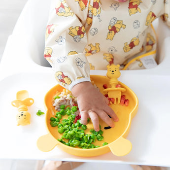 Baby practicing self-feeding with peas on a bear plate, wearing Bumkins Winnie Loves Hunny sleeved bib.