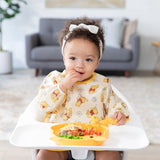 A toddler in a Bumkins Sleeved Bib: Winnie Loves Hunny sits in a high chair, eating from a yellow plate, ideal for baby-led weaning.