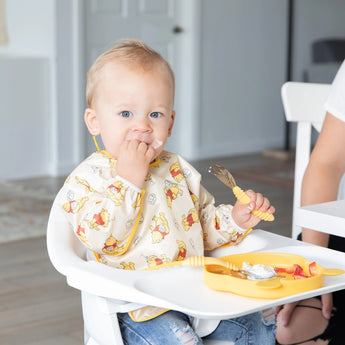 A baby in a high chair, wearing the Bumkins Sleeved Bib: Winnie Loves Hunny, enjoys baby-led weaning with a yellow fork and plate.