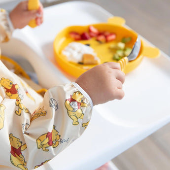 A child in a Winnie Loves Hunny sleeved bib by Bumkins holds a fork and spoon over a yellow plate for baby-led weaning.