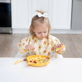 A toddler in a Bumkins Winnie Loves Hunny sleeved bib uses a fork to eat from a yellow bowl, exploring baby-led weaning at the table.