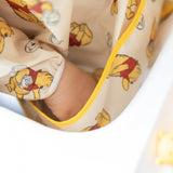 Close-up of childs hand in Bumkins Sleeved Bib: Winnie Loves Hunny, featuring yellow cartoon bears with red shirts and yellow piping.