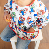 Child in a Bumkins Sleeved Bib: Super Mario™ Classic, sitting on a stool holding a cookie, with the bib catching crumbs.