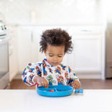 A toddler in a Bumkins Sleeved Bib: Super Mario™ Classic enjoys food from a blue plate at the kitchen table.