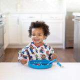 A toddler smiles at the table, wearing a Bumkins Sleeved Bib: Super Mario™ Classic, eating from a blue plate in a bright kitchen.