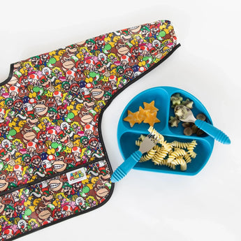 Bumkins Sleeved Bib: Super Mario™ Mashup beside a blue divided plate with pasta, star-cut cheese, broccoli, and utensils.