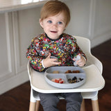 A toddler in a patterned shirt sits in a high chair with snacks on a white tray, wearing the Bumkins Sleeved Bib: Super Mario™ Mashup.