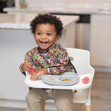 A happy toddler in a Bumkins Sleeved Bib: Super Mario™ Mashup holds a pink toy, sitting in a high chair with food on the tray.