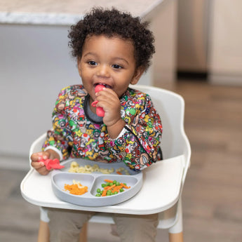 A toddler in a Bumkins Super Mario™ Mashup bib eats happily from a plate in a high chair, holding a spoon.