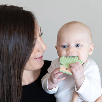 A woman holds her baby happily chewing on Bumkins cactus-shaped Silicone Teether for soothing relief.