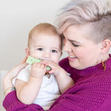 A person with short hair smiles joyfully as the baby finds comfort with a Bumkins Silicone Teether: Cactus.