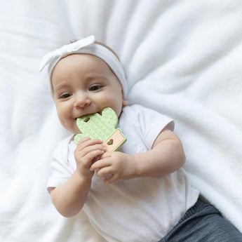 A baby smiles on a soft surface, wearing a headband and holding Bumkins Silicone Teether: Cactus for teething relief.