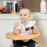 A baby holds a retro game controller toy and the Bumkins Silicone Teether: Game Boy™ while in the kitchen high chair.