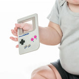 A baby in a gray outfit holds a Bumkins Silicone Teether: Game Boy™, against a white background, ideal for teething relief.