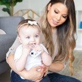 A woman holds a baby happily chewing on a Bumkins Silicone Teether: Game Boy™ while sitting on the cozy living room floor.