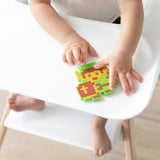 A child holds a Bumkins Zelda™ Link silicone teether with green, yellow, and red colors on a white high chair tray.